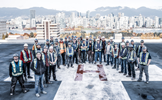 A group of Modern Niagara employees in safety gear on a helicopter pad on the roof of the building with Downtown Vancouver in the backdrop